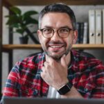 Head shot portrait of positive freelance businessman in eyeglasses sitting at home office